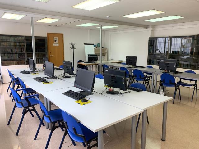 Aula del Punt Òmnia Casa del Mar de Vilanova i la Geltrú, amb diverses taules blanques disposades en forma de U i equipades amb ordinadors de sobretaula, teclats i ratolins. Hi ha cadires blaves, una pissarra blanca, una porta amb cartell, un penja-robes i una prestatgeria amb carpetes.