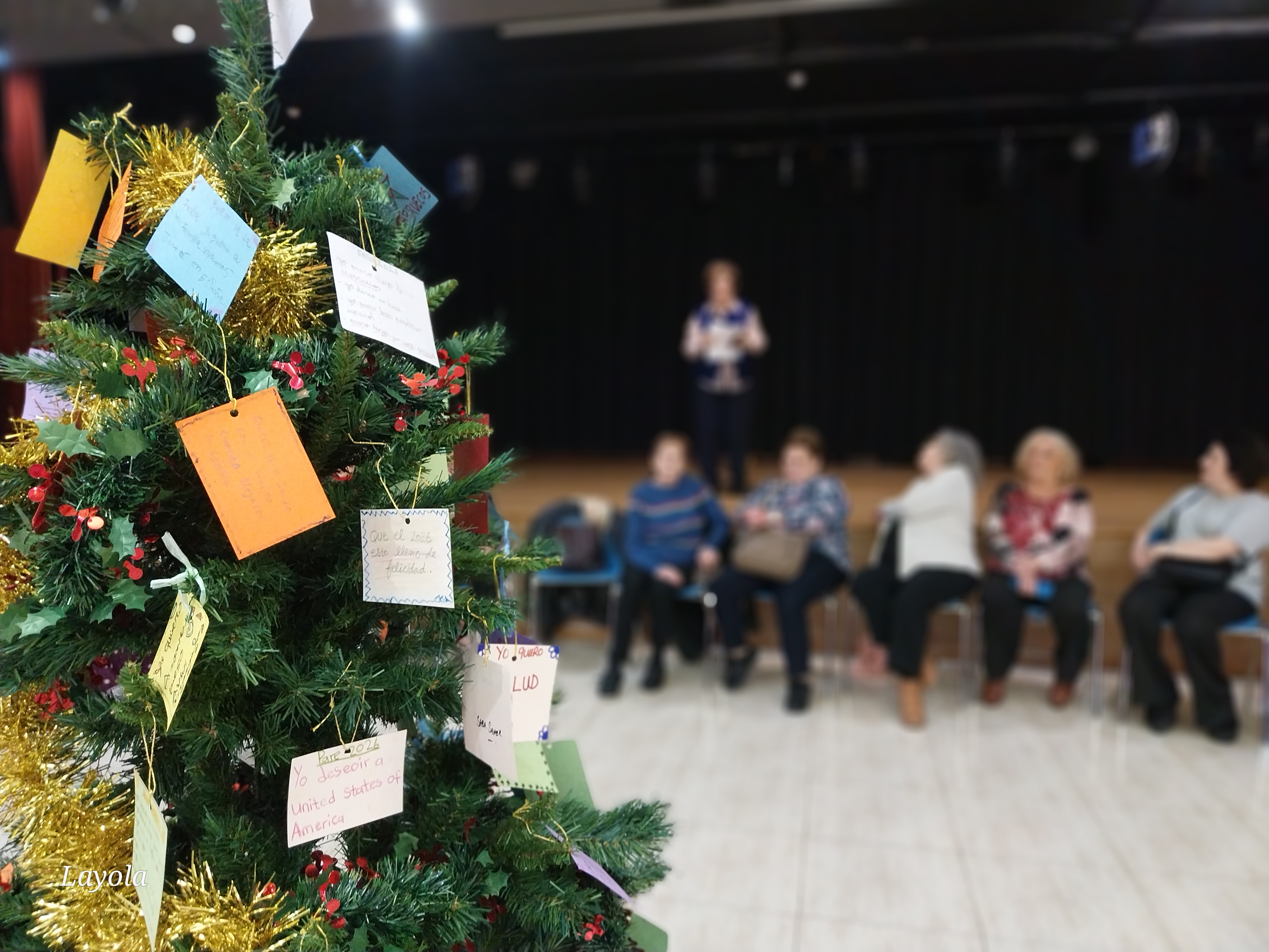 Imatge d'un arbre de Nadal on pengen papers de colors petits. Al fons, desenfocat, es veuen sis dones que xerren entre elles. 