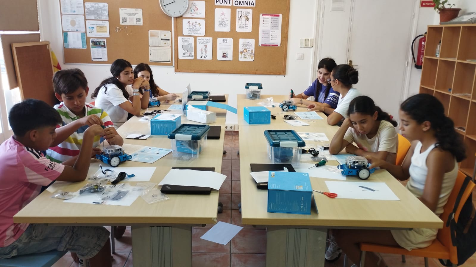 Un grup de 8 infants, majoritàriament nenes, s'ubiquen al voltant d'una taula mentre manipulen robots educatius. 