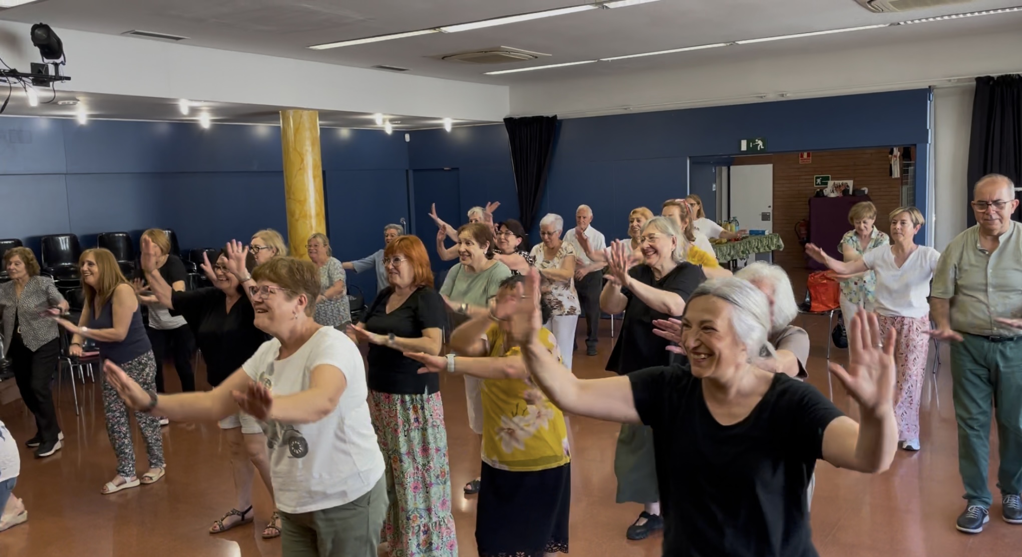  Grup de persones grans, majoritàriament dones, ballant i somrient en una sala espaiosa durant una activitat col·lectiva. Tothom té els braços aixecats i segueix una coreografia, transmetent alegria i complicitat.