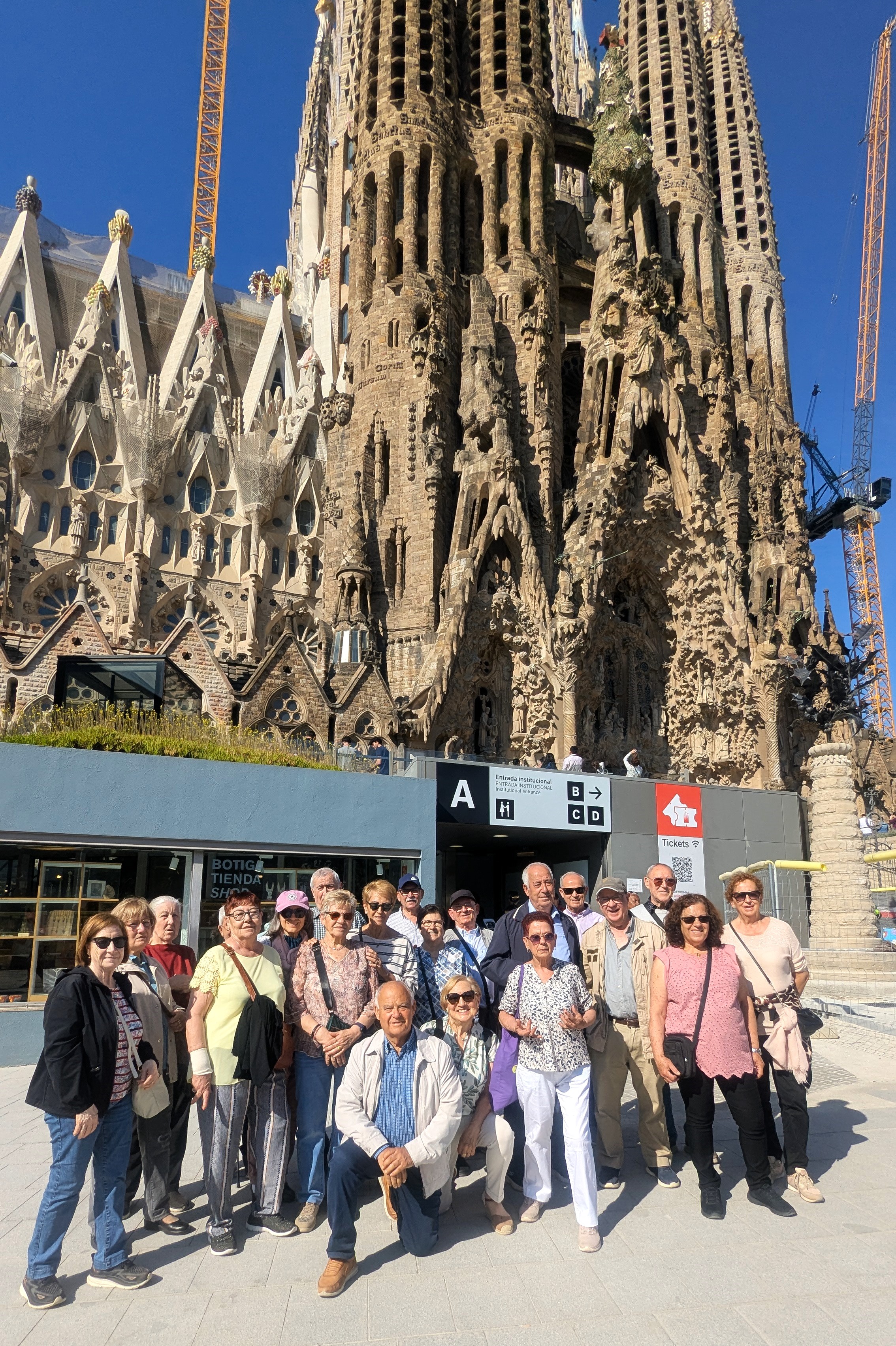 Un grup de gent gran de 20 persones posen davant de la Sagrada Família. És un dia assolellat i molts d'ells duen gorres i ulleres de sol. 
