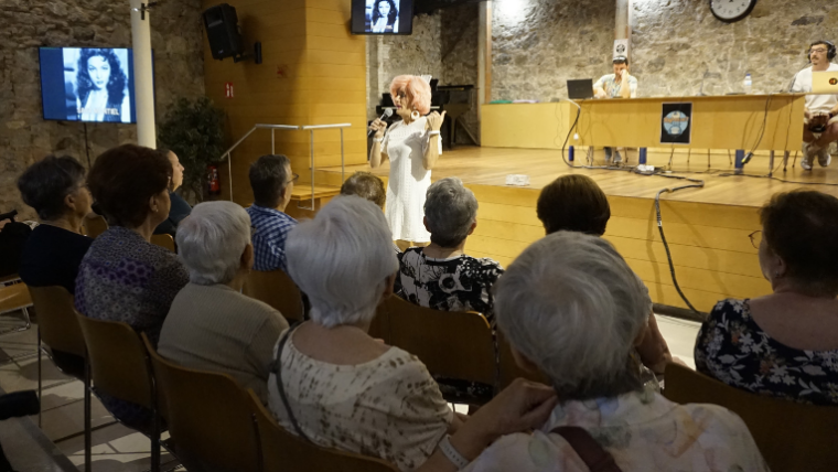Aquí tens una proposta de **text alternatiu (text alt)** per a la imatge, coherent amb el context que m’has facilitat:  **Text alt:** *Un grup de persones grans seuen a la sala polivalent d’un equipament comunitari del barri del Carmel, participant en una activitat del projecte Refresca’t. Al fons, la presentadora Brigitta Lamoure condueix una dinàmica col·lectiva amb micròfon a la mà, mentre el públic escolta atent i participa. La imatge reflecteix un ambient proper i intergeneracional, amb la projecció d’