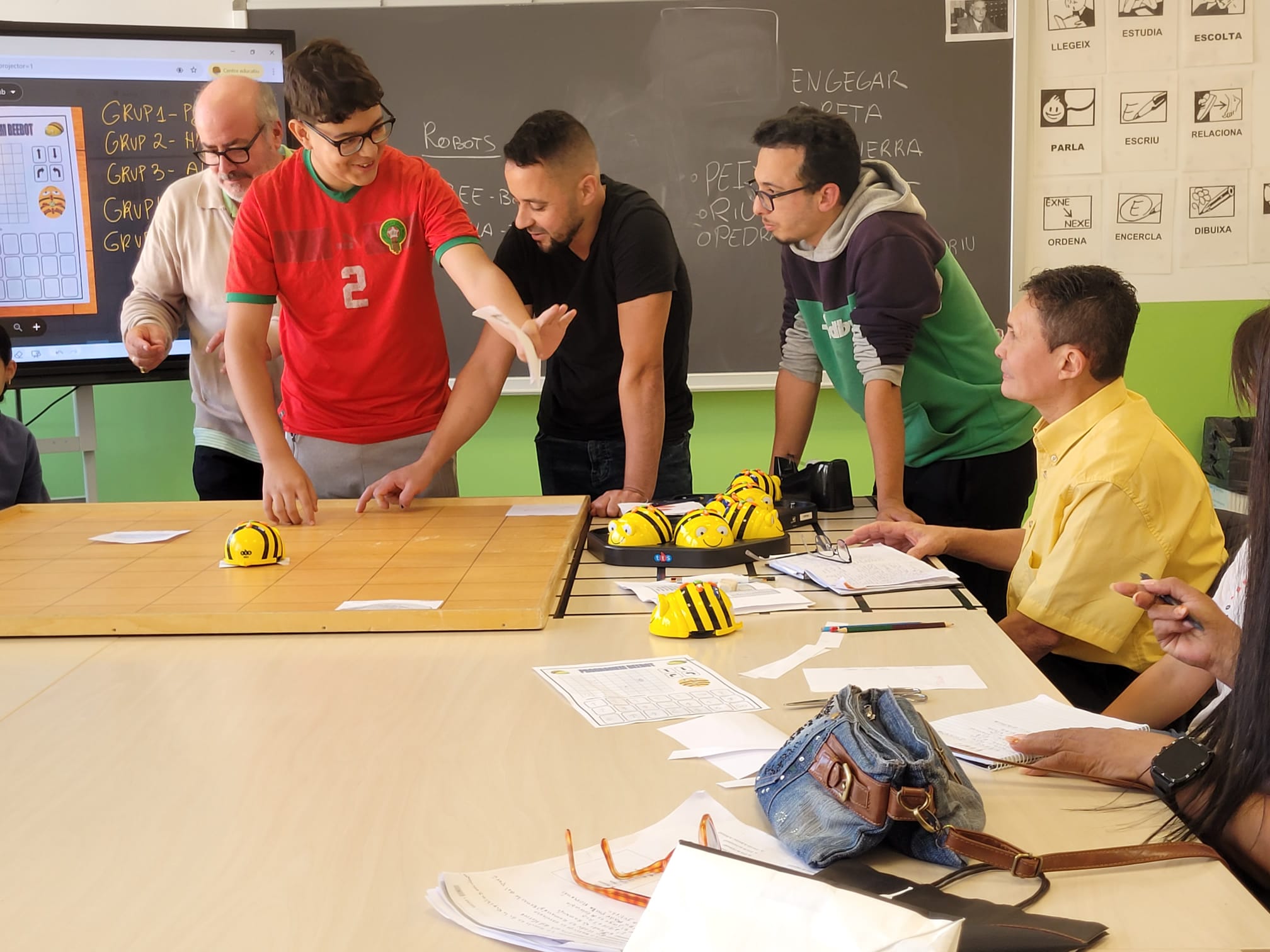 Participants de diferents edats en un taller de robòtica intergeneracional al Punt Òmnia Eudald Graells de Ripoll, programant i movent petits robots educatius sobre una taula, mentre prenen apunts i comparteixen experiències.