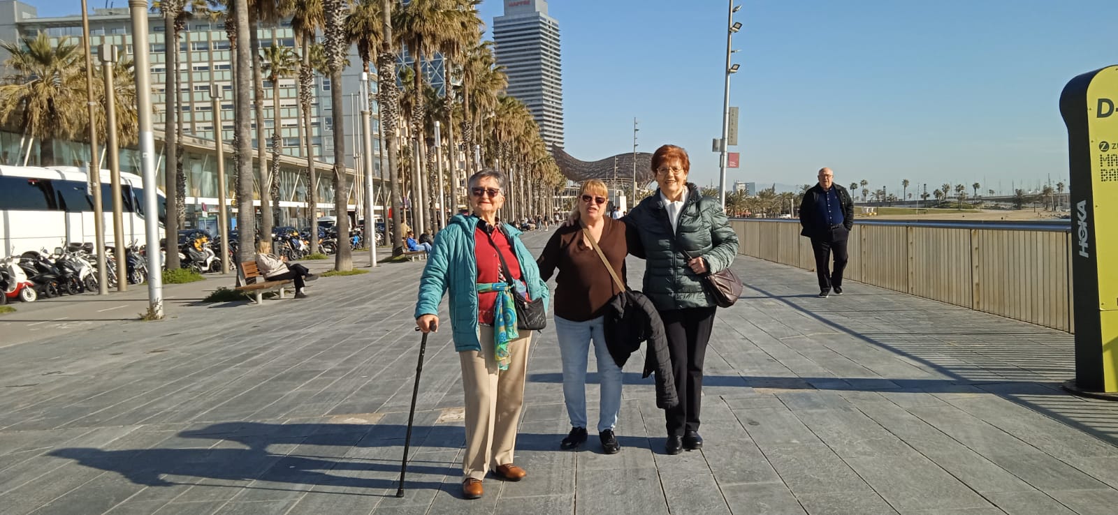 Tres dones grans somriuen mirant a càmera, situades en un passeig al costat d'una platja. 