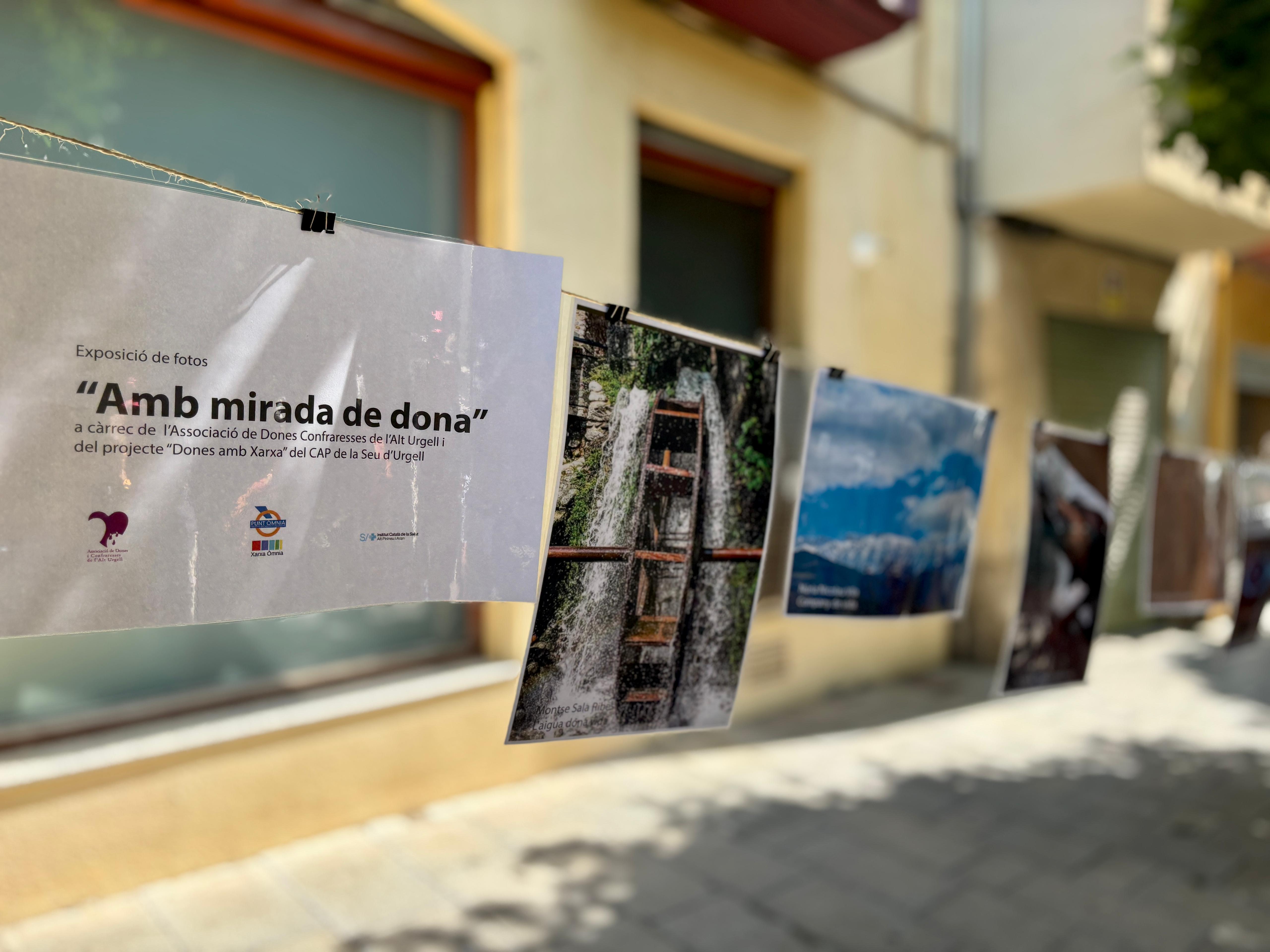 Imatge de l'exposició "Amb mirada de dona". Apareix un cartell amb el nom de l'exposició imprès i es veuen de fons algunes de les fotografies seleccionades. Imatge de l'exposició "Amb mirada de dona". Apareix un cartell amb el nom de l'exposició imprès i es veuen de fons algunes de les fotografies seleccionades.