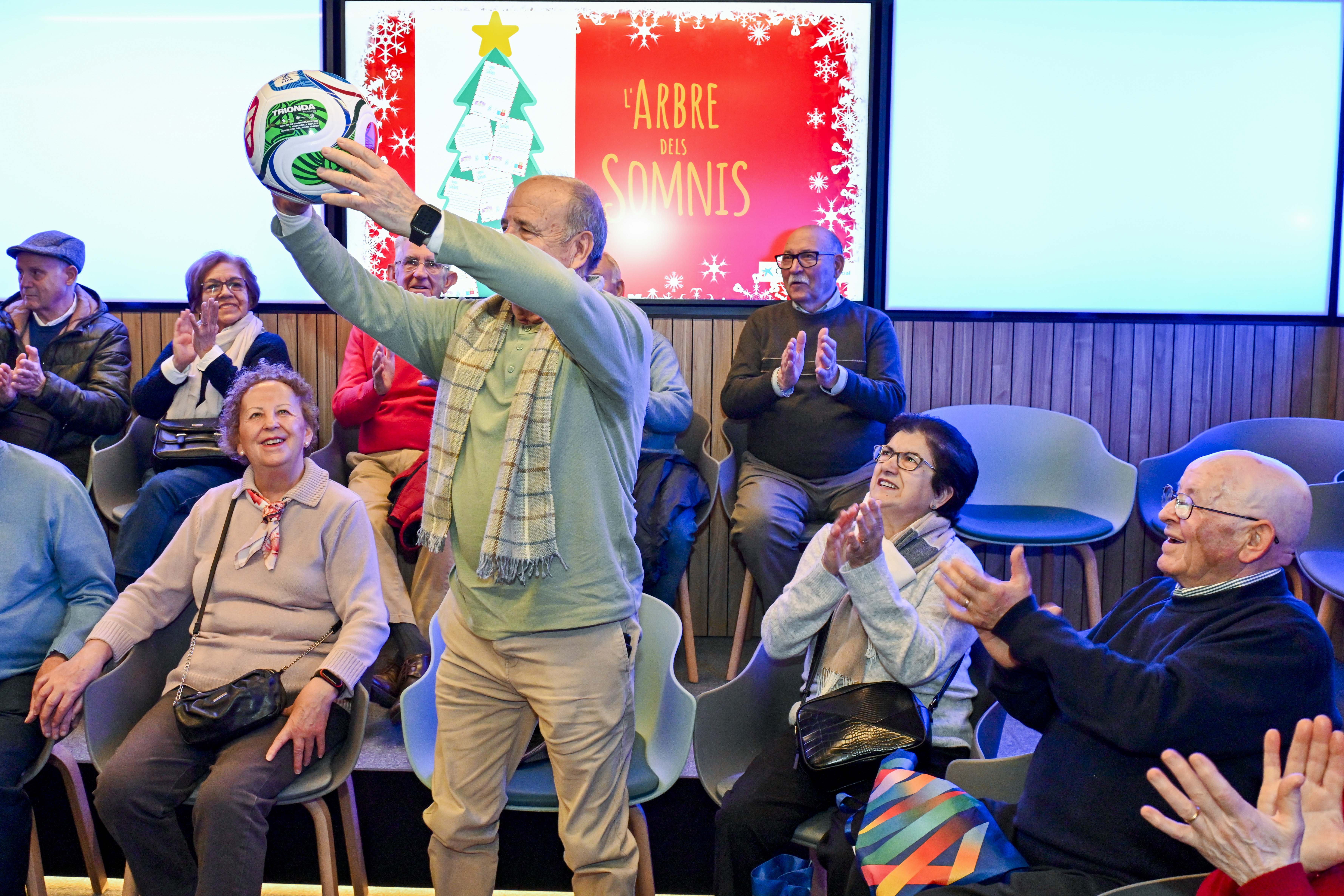 Un grup de persones grans assegudes a cadires somriuen i aplaudeixen. Al centre, un home de peu agafa una pilota de futbol. Al fons, es llegeix sobre una pantalla "L'arbre dels somnis".