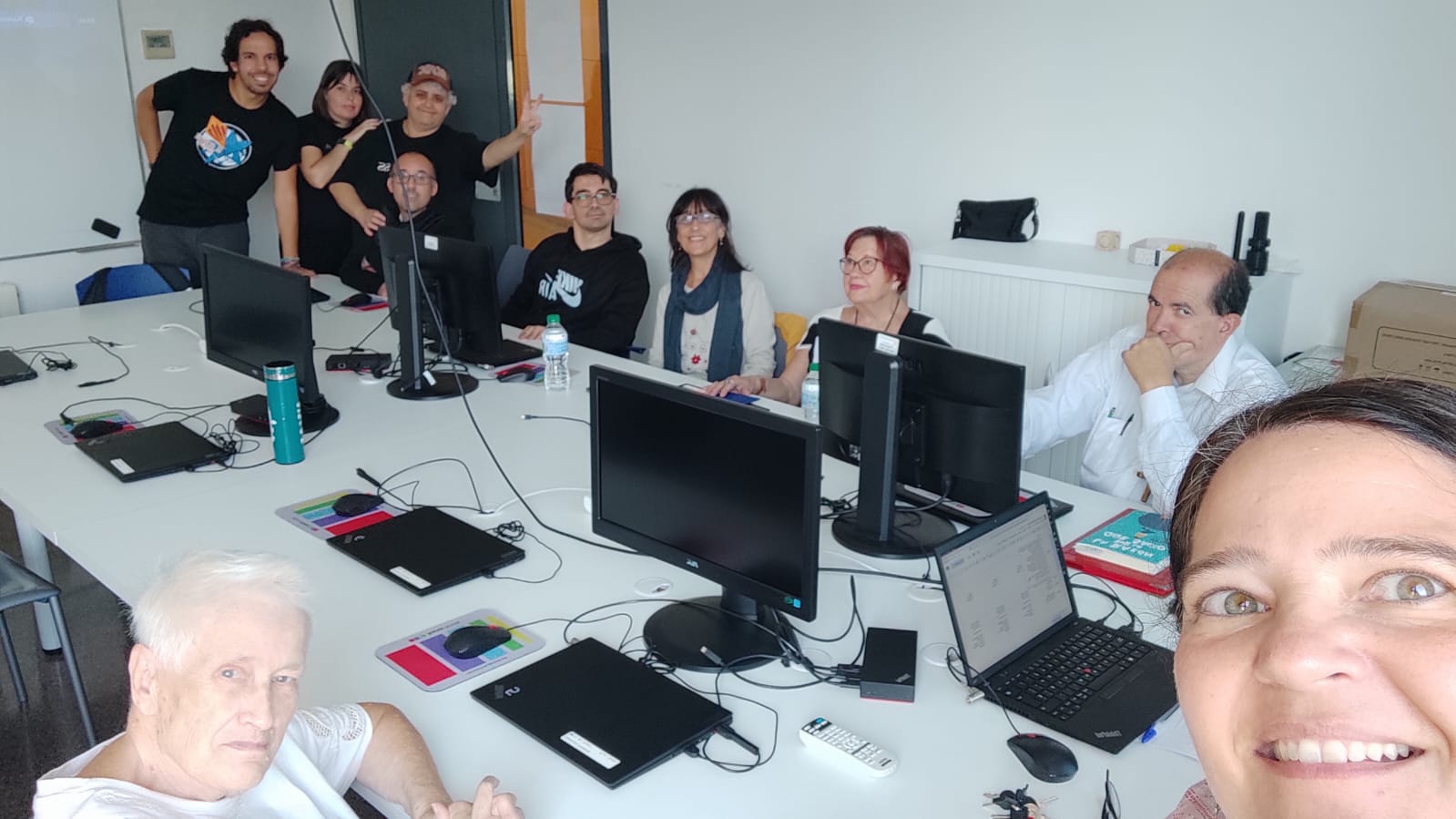 Fotografia d’un grup de deu persones en una aula amb ordinadors. La majoria estan assegudes darrere d’una taula llarga amb monitors i portàtils, mentre dues persones estan dempeus al fons. Al primer pla, una dona fa un selfie somrient.