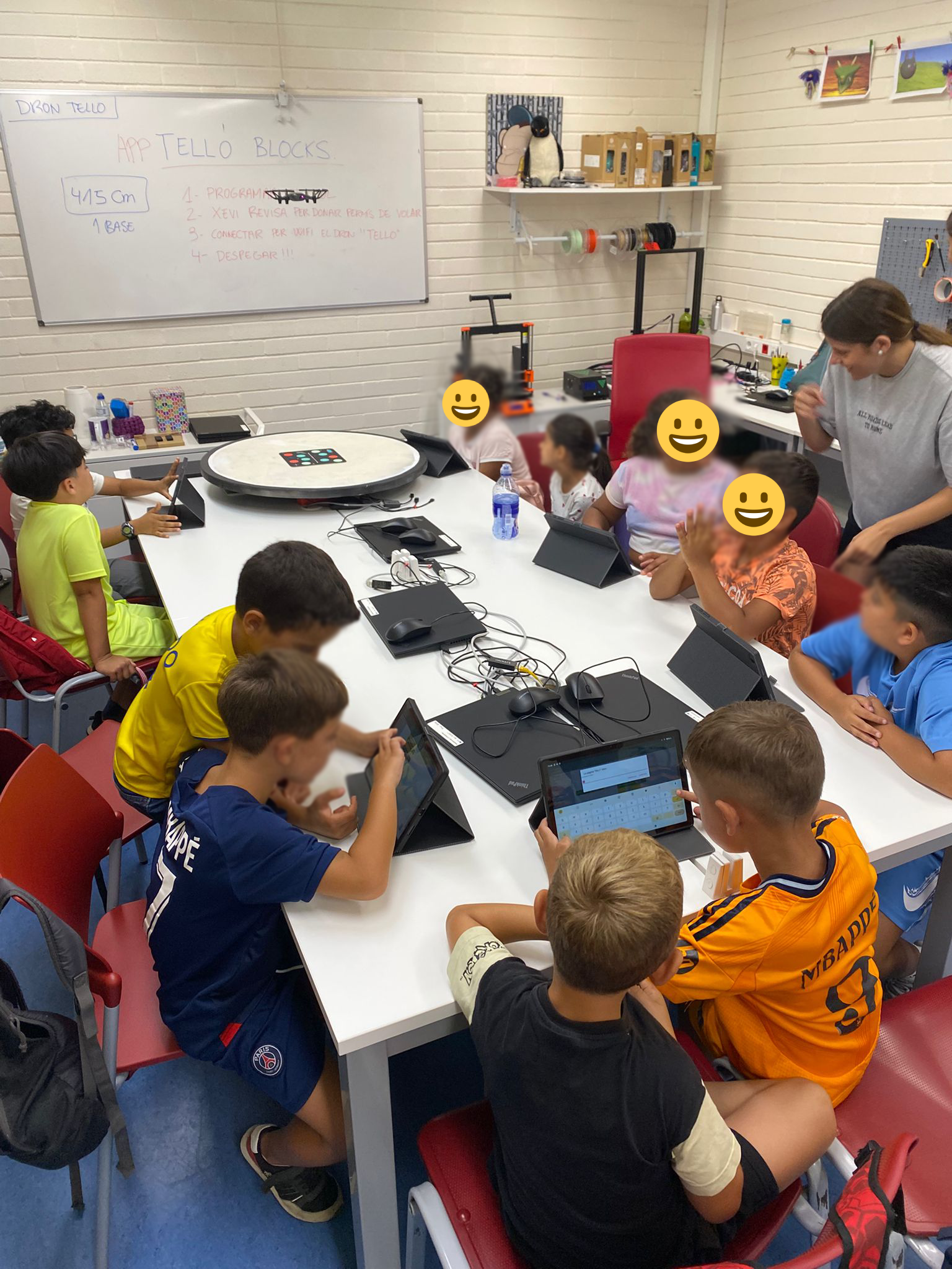 Infants asseguts al voltant d’una taula blanca en una aula equipada amb ordinadors i tauletes, participant en un taller de programació amb l'app Tello Blocks per controlar un dron. A la pissarra es poden llegir instruccions bàsiques de vol. Una dinamitzadora acompanya el grup.