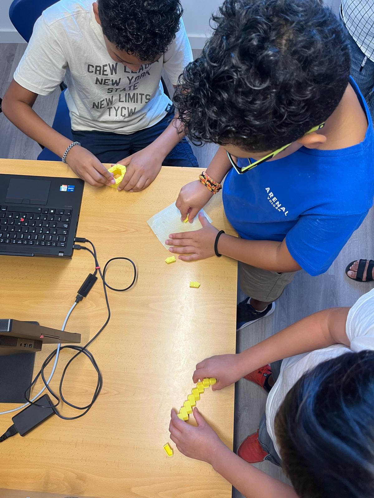 Tres joves treballen en grup al voltant d’una taula, manipulant petites peces grogues impreses en 3D al costat d’un ordinador portàtil.