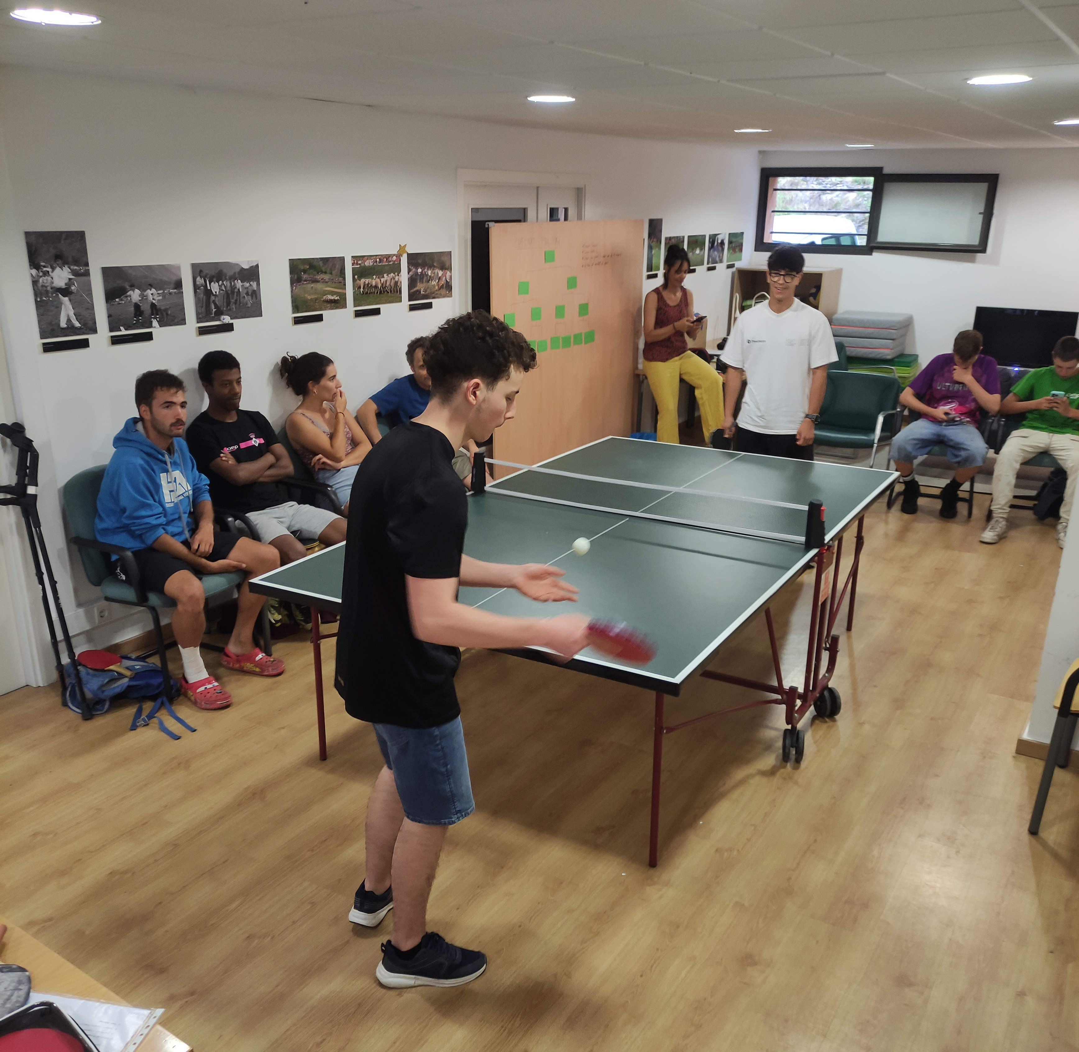 Grup de joves jugant a tennis taula en una sala amb parquet i parets decorades amb fotografies. Diversos nois i noies seuen al voltant observant el partit, mentre dos nois juguen a la taula de ping-pong. Al fons, una monitora pren notes i altres participants miren el mòbil o conversen.