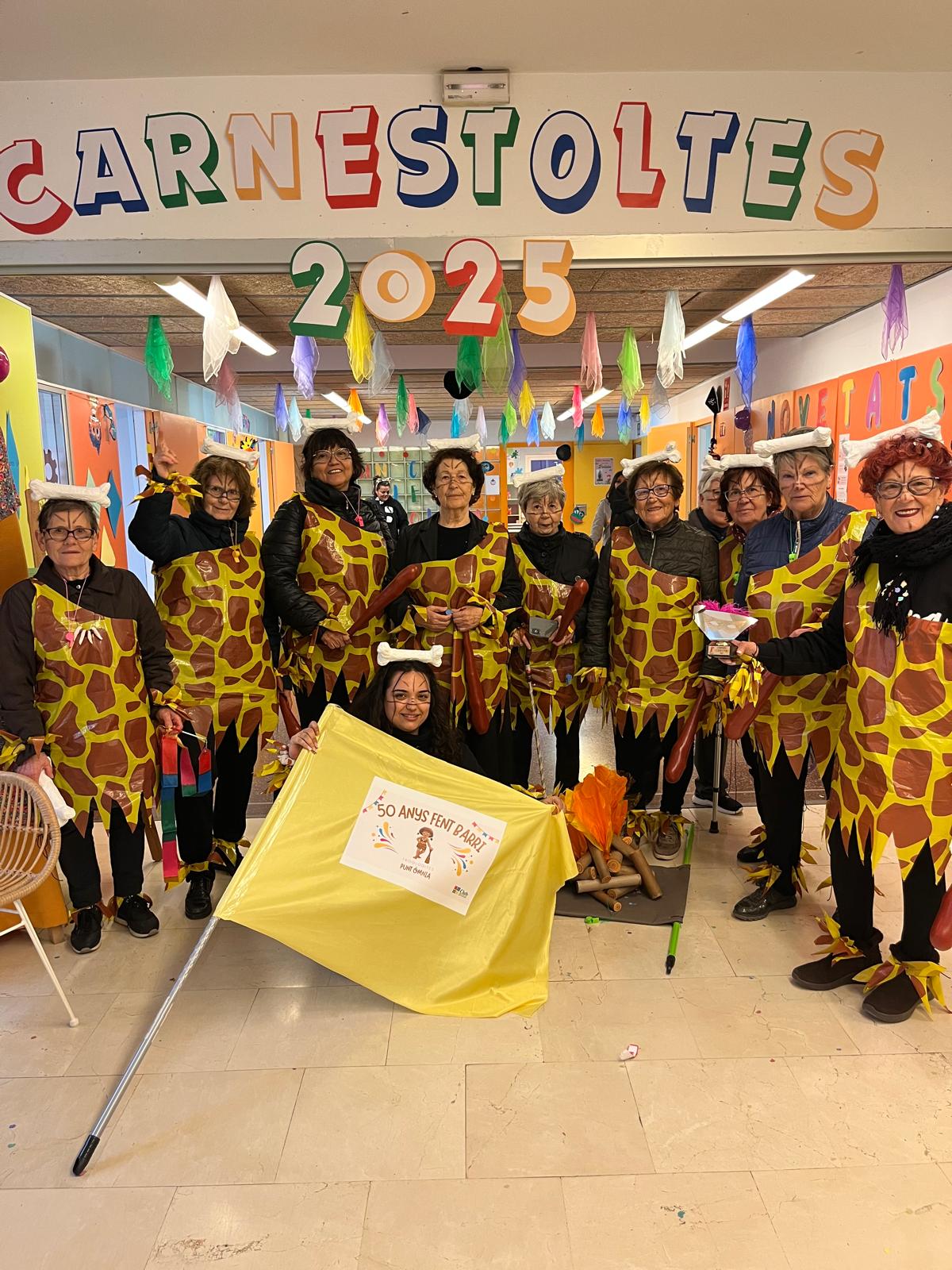 Un grup de dones grans disfressades de trogodites amb un os al cap posen per la fotografia sota un cartell de colors on es llegeix "Carnaval 2025".