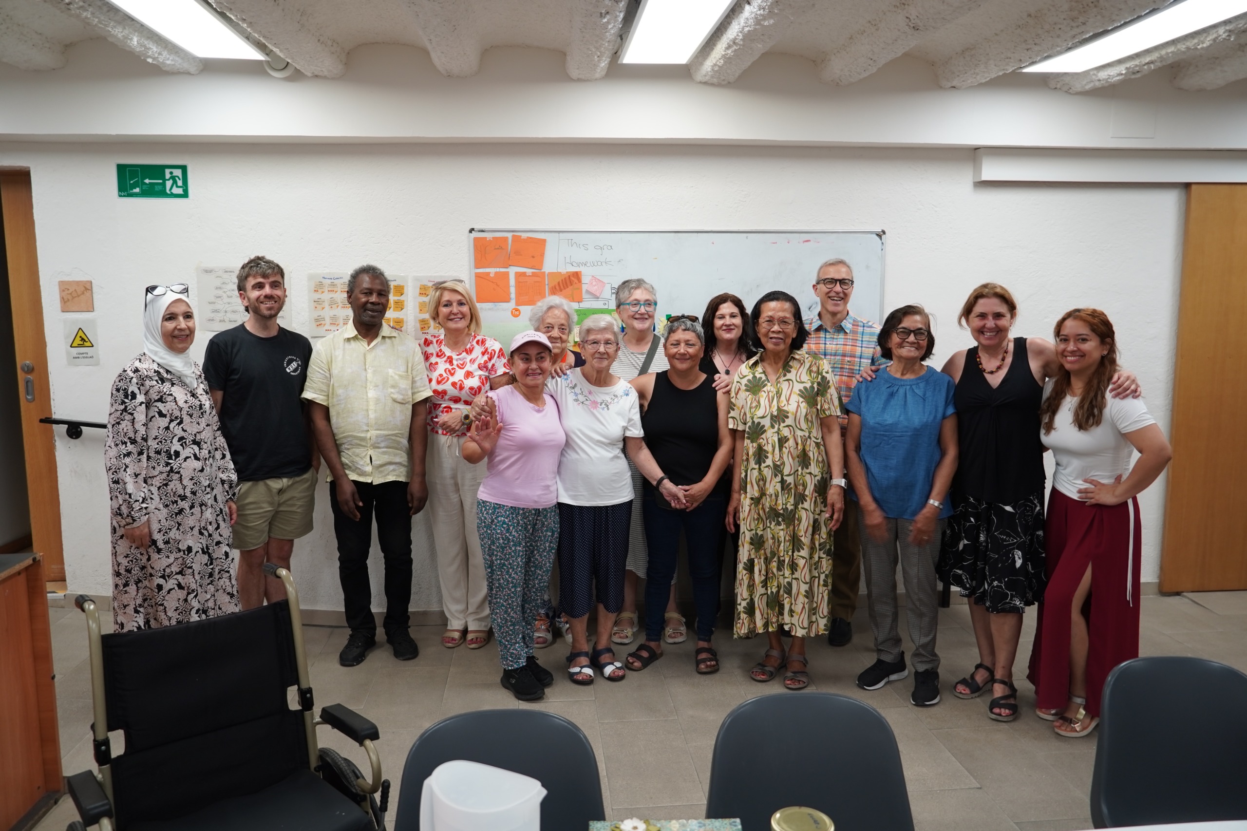 Foto de grup de les participants i dinamitzadores del Punt Òmnia del Raval durant la cloenda del projecte de capacitació digital per a persones grans. Es veuen diverses persones grans i membres de l’equip, somrients, drets davant d’una pissarra amb cartells de colors i missatges. Al primer pla hi ha una cadira de rodes i part del mobiliari de l’aula.