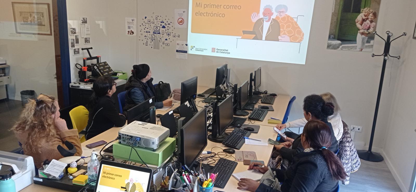 Grup de dones assegudes a una aula equipada amb ordinadors que miren una pantalla on es projecta la presentació d'una formació sobre correu electrònic.