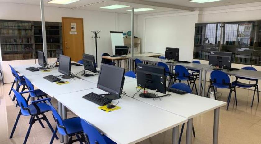 Aula del Punt Òmnia Casa del Mar de Vilanova i la Geltrú, amb diverses taules blanques disposades en forma de U i equipades amb ordinadors de sobretaula, teclats i ratolins. Hi ha cadires blaves, una pissarra blanca, una porta amb cartell, un penja-robes i una prestatgeria amb carpetes.