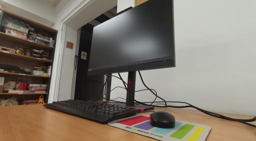 Monitor d’ordinador encès sobre una taula de fusta clara, amb teclat i ratolí col·locats a davant. El ratolí està sobre una catifa de ratolí amb el logotip i els colors de la Xarxa Òmnia. 