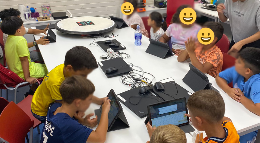 Infants asseguts al voltant d’una taula blanca en una aula equipada amb ordinadors i tauletes, participant en un taller de programació amb l'app Tello Blocks per controlar un dron. A la pissarra es poden llegir instruccions bàsiques de vol. Una dinamitzadora acompanya el grup.