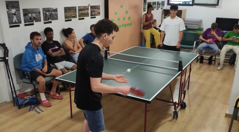 Grup de joves jugant a tennis taula en una sala amb parquet i parets decorades amb fotografies. Diversos nois i noies seuen al voltant observant el partit, mentre dos nois juguen a la taula de ping-pong. Al fons, una monitora pren notes i altres participants miren el mòbil o conversen.