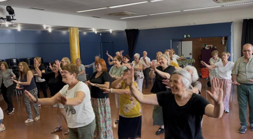  Grup de persones grans, majoritàriament dones, ballant i somrient en una sala espaiosa durant una activitat col·lectiva. Tothom té els braços aixecats i segueix una coreografia, transmetent alegria i complicitat.