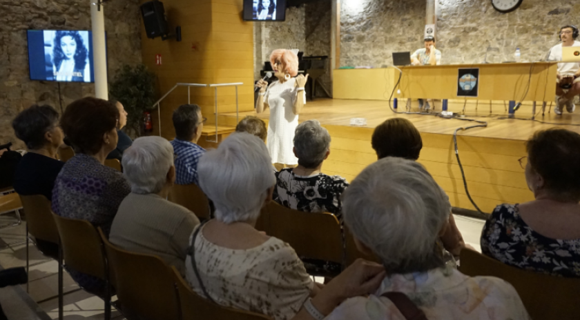 Aquí tens una proposta de **text alternatiu (text alt)** per a la imatge, coherent amb el context que m’has facilitat:  **Text alt:** *Un grup de persones grans seuen a la sala polivalent d’un equipament comunitari del barri del Carmel, participant en una activitat del projecte Refresca’t. Al fons, la presentadora Brigitta Lamoure condueix una dinàmica col·lectiva amb micròfon a la mà, mentre el públic escolta atent i participa. La imatge reflecteix un ambient proper i intergeneracional, amb la projecció d’