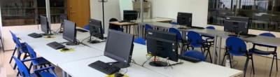 Aula del Punt Òmnia Casa del Mar de Vilanova i la Geltrú, amb diverses taules blanques disposades en forma de U i equipades amb ordinadors de sobretaula, teclats i ratolins. Hi ha cadires blaves, una pissarra blanca, una porta amb cartell, un penja-robes i una prestatgeria amb carpetes.