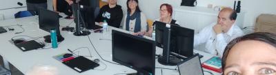 Fotografia d’un grup de deu persones en una aula amb ordinadors. La majoria estan assegudes darrere d’una taula llarga amb monitors i portàtils, mentre dues persones estan dempeus al fons. Al primer pla, una dona fa un selfie somrient.