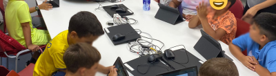 Infants asseguts al voltant d’una taula blanca en una aula equipada amb ordinadors i tauletes, participant en un taller de programació amb l'app Tello Blocks per controlar un dron. A la pissarra es poden llegir instruccions bàsiques de vol. Una dinamitzadora acompanya el grup.