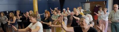  Grup de persones grans, majoritàriament dones, ballant i somrient en una sala espaiosa durant una activitat col·lectiva. Tothom té els braços aixecats i segueix una coreografia, transmetent alegria i complicitat.